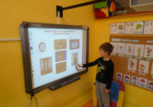 Chłopiec stoi obok tablicy interaktywnej, w prawej ręce trzyma wskaźnik, którym pokazuje zegar wahadłowy.